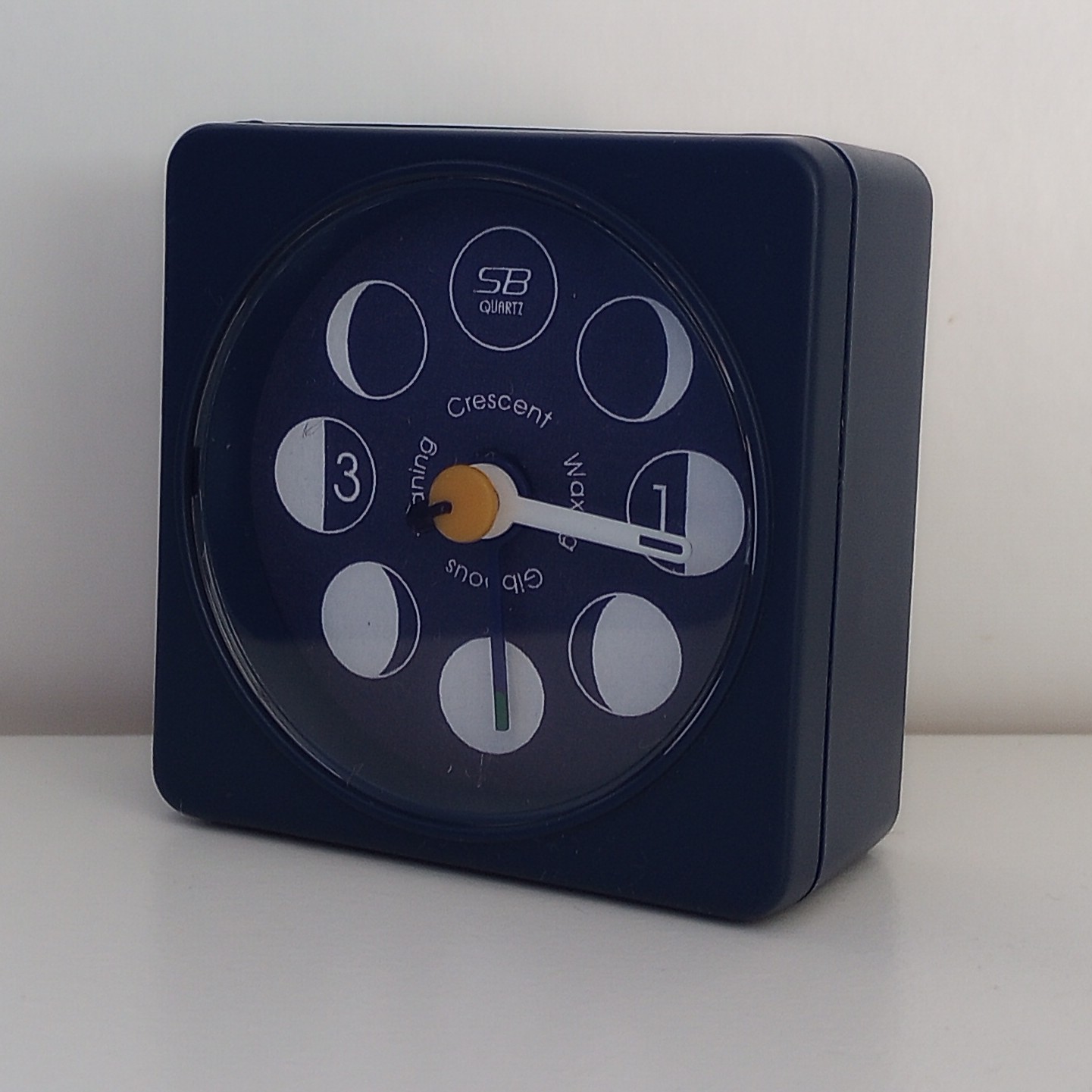 A small dark blue alarm clock, with the face showing the phases of the moon, and a single hand (currently pointing to the first quarter, just past Waxing Gibbous).